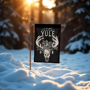 May include: A black garden flag with a white illustration of a deer skull with antlers and a wreath of holly and berries. The text "a bright and peaceful YULE" is above the skull.