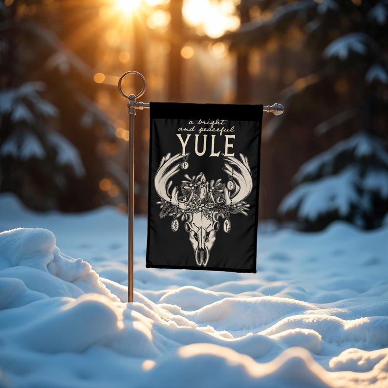 May include: A black garden flag with a white illustration of a deer skull with antlers and a wreath of holly and berries. The text "a bright and peaceful YULE" is above the skull.