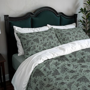 May include: A bed with a dark green headboard and white bedding. The duvet cover and pillowcases feature a repeating pattern of various moths and insects in black on a muted green background. The bed is made up with white pillows and a white duvet.