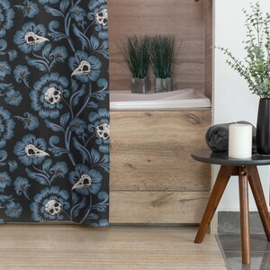 Puede incluir: Una cortina de ducha con un patr&oacute;n floral azul oscuro con calaveras blancas y hojas. La cortina est&aacute; colgada en un ba&ntilde;o con una ba&ntilde;era de madera y una mesa auxiliar con una vela y un jarr&oacute;n de plantas verdes.