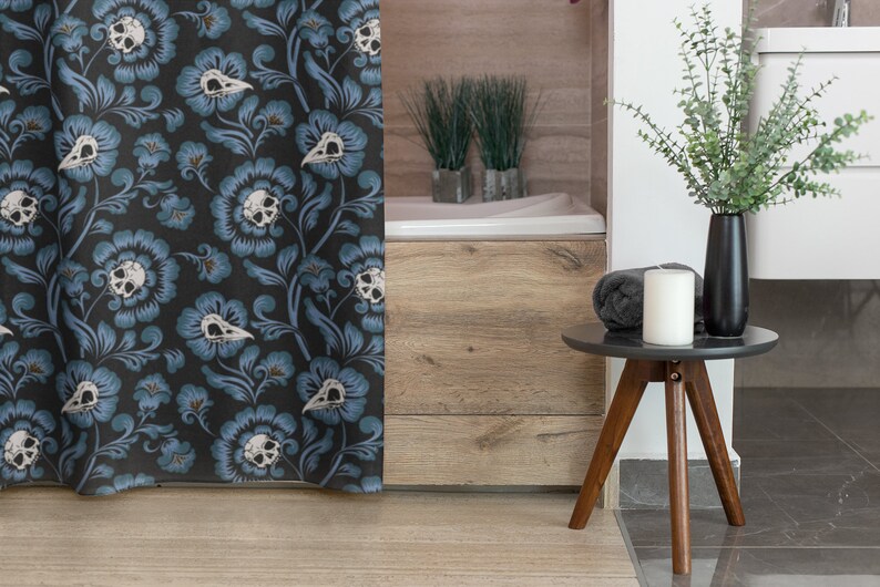 Puede incluir: Una cortina de ducha con un patr&oacute;n floral azul oscuro con calaveras blancas y hojas. La cortina est&aacute; colgada en un ba&ntilde;o con una ba&ntilde;era de madera y una mesa auxiliar con una vela y un jarr&oacute;n de plantas verdes.