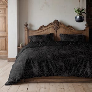 Op de afbeelding: Zwarte beddengoedset met een spinnenwebontwerp. De set bevat een dekbedovertrek en twee kussenslopen. Het bed heeft een houten hoofdbord en een houten bedframe. De kamer heeft een neutraal kleurenpalet.