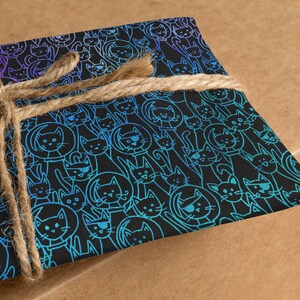K&ouml;nnte beinhalten: Ein Geschenk, das in blau und violett gemustertes Papier mit schwarzem Hintergrund eingewickelt ist. Das Papier zeigt ein sich wiederholendes Muster von Katzen mit verschiedenen Gesichtsausdr&uuml;cken. Das Geschenk ist mit einer braunen Schnur gebunden.