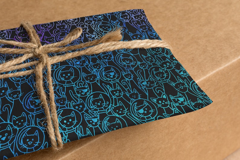 K&ouml;nnte beinhalten: Ein Geschenk, das in blau und violett gemustertes Papier mit schwarzem Hintergrund eingewickelt ist. Das Papier zeigt ein sich wiederholendes Muster von Katzen mit verschiedenen Gesichtsausdr&uuml;cken. Das Geschenk ist mit einer braunen Schnur gebunden.