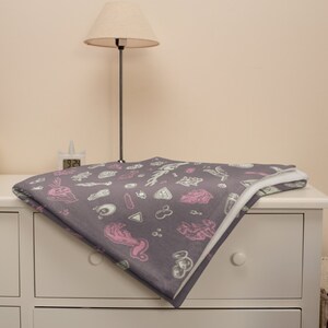 May include: A grey throw blanket with a white and pink pattern of various occult symbols. The blanket is folded and placed on a white dresser.