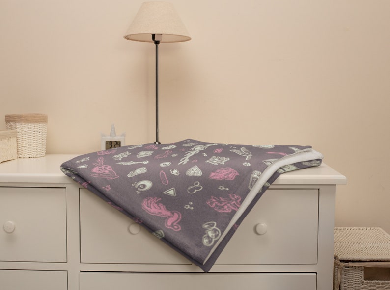 May include: A grey throw blanket with a white and pink pattern of various occult symbols. The blanket is folded and placed on a white dresser.