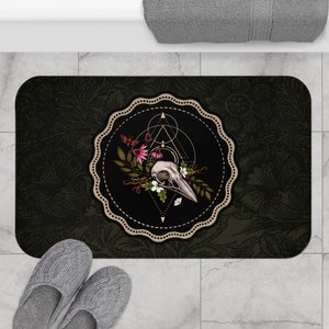 Puede incluir: Una alfombra de baño negra y dorada con un patrón floral y una calavera con un pico de pájaro en el centro. La calavera está rodeada por un círculo de hojas y flores.