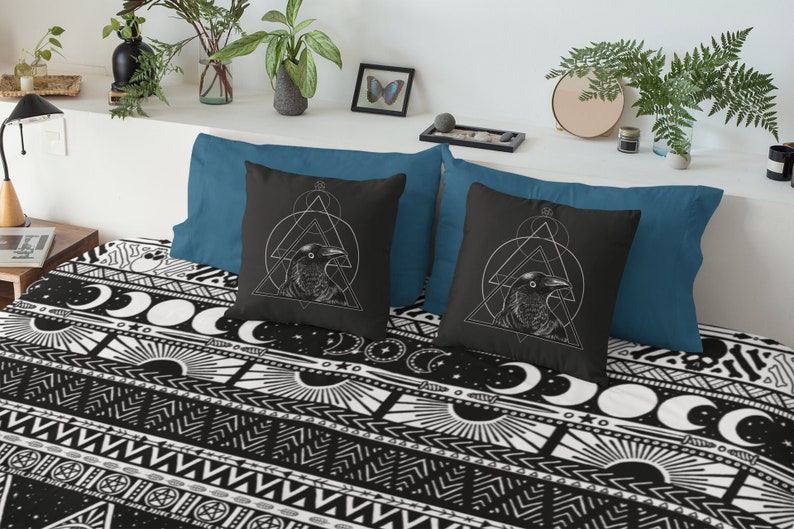 May include: Two black throw pillows with white geometric designs and a raven illustration. The pillows are on a bed with a black and white patterned bedspread.