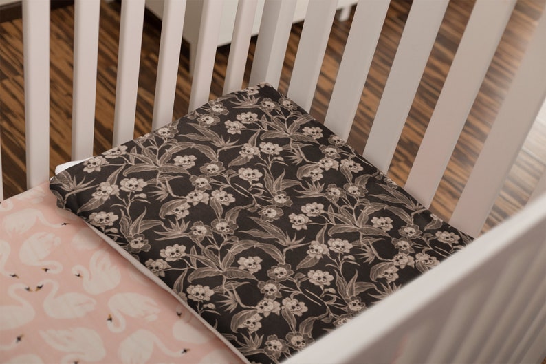 May include: A white crib with a black and white skull and floral patterned fitted sheet and a pink fitted sheet with white swans.