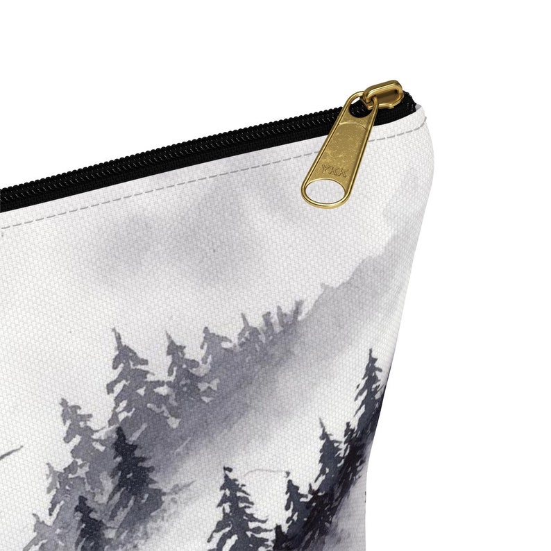 May include: A white zippered pouch with a black zipper and a gold zipper pull. The pouch features a watercolor-style design of a misty forest with black trees.