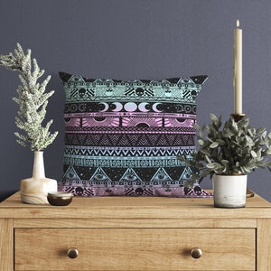 May include: A black and pink throw pillow with a geometric pattern featuring moons, stars, and eyes. The pillow is on a wooden table with a candle and a plant.