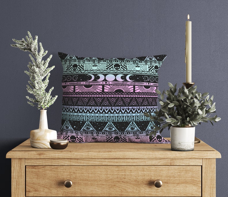 May include: A black and pink throw pillow with a geometric pattern featuring moons, stars, and eyes. The pillow is on a wooden table with a candle and a plant.