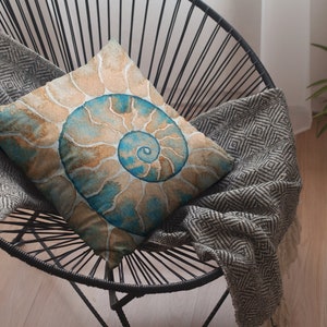 May include: A decorative throw pillow with a watercolor design of a nautilus shell in shades of blue, brown, and beige. The pillow is sitting on a black chair with a gray and white patterned throw blanket.