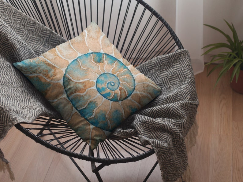 May include: A decorative throw pillow with a watercolor design of a nautilus shell in shades of blue, brown, and beige. The pillow is sitting on a black chair with a gray and white patterned throw blanket.