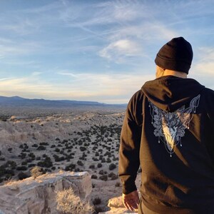 May include: Black zip-up hoodie with a detailed white graphic of a skull and floral design on the back. The person is wearing a black beanie and is overlooking a desert landscape under a blue sky.