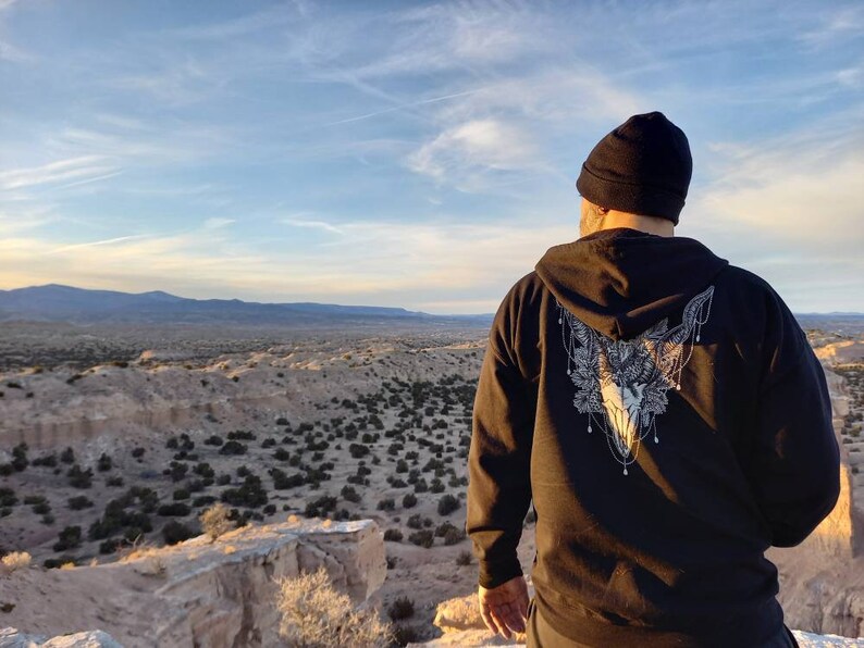 May include: Black zip-up hoodie with a detailed white graphic of a skull and floral design on the back. The person is wearing a black beanie and is overlooking a desert landscape under a blue sky.