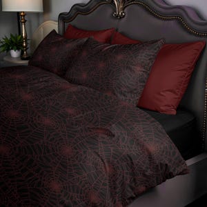 以下が含まれることがあります： 赤色のクモの巣模様の黒い掛け布団と枕カバーがセットされたベッド。2つの無地の赤い枕がヘッドボードに寄りかかっています。ベッドは整えられており、すぐに使用できます。