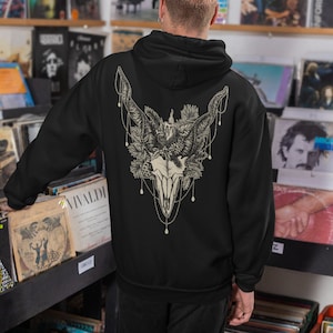 May include: Black hoodie featuring a detailed graphic of a skull with horns, floral elements, and beaded accents on the back. The design is in shades of cream and beige. The hoodie is a casual garment.