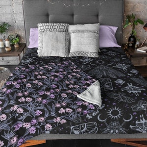 Throw gothic blanket on bed with decorative pillows, fairy lights and purple pillows. Blanket is gothic with pink, purple and blue skull flowers.