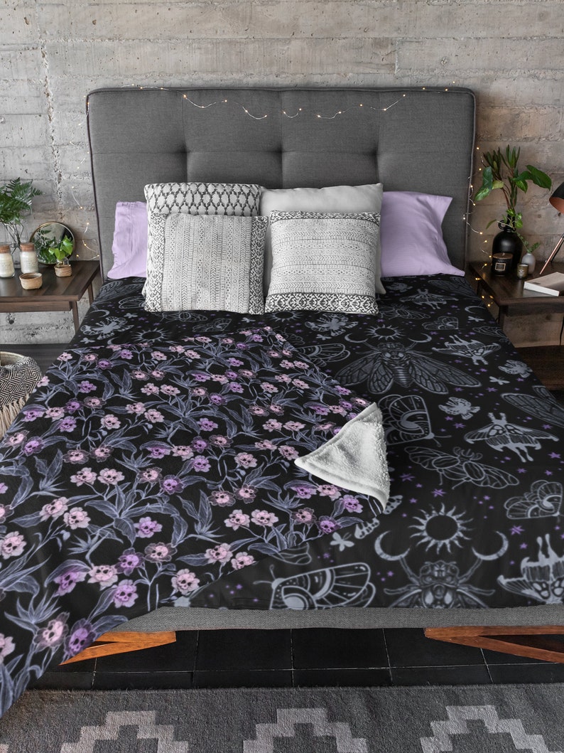 Throw gothic blanket on bed with decorative pillows, fairy lights and purple pillows. Blanket is gothic with pink, purple and blue skull flowers.