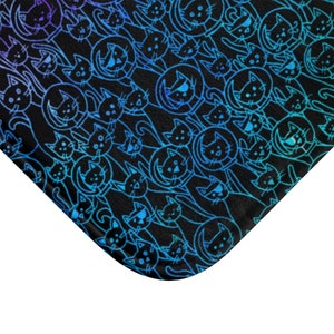 以下が含まれることがあります： 黒いバスマット。青とターコイズの猫の顔が繰り返されるパターン。猫の顔は青とターコイズのグラデーションで縁取られ、背景は黒です。マットは角が丸くなっています。