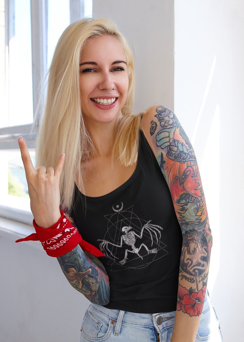 May include: A black tank top with a white graphic of a bat skeleton inside a geometric design. The tank top is being worn by a person with tattoos on their arms. The person is wearing a red bandana on their wrist and is making a rock and roll hand gesture.