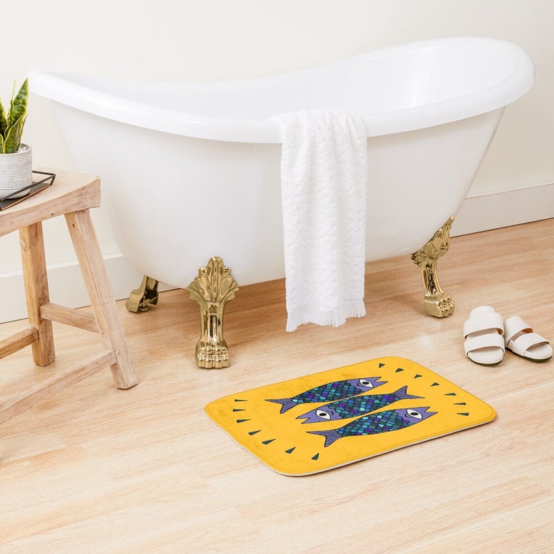 Puede incluir: Una alfombra de ba&ntilde;o amarilla con un dise&ntilde;o de tres peces azules y morados con ojos negros. Los peces nadan en fila y tienen un fondo amarillo.