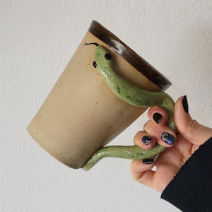 May include: A beige ceramic mug with a brown rim and a green snake handle. The snake handle is detailed with black eyes and a forked tongue. The mug has a speckled texture and is held by a person.