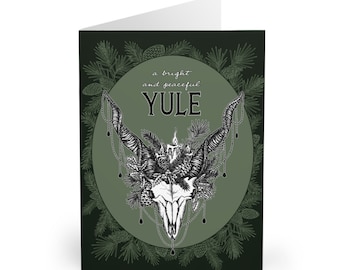 Könnte beinhalten: Eine grüne Karte mit einer Schwarzweißillustration eines Schädels mit Geweih und einer Kerze darauf. Der Schädel ist von Kiefernästen und Tannenzapfen umgeben. Der Text "a bright and peaceful YULE" ist in Schwarz auf einem grünen Kreis geschrieben.