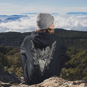 May include: A black hoodie with a detailed graphic of a skull with antlers, pine needles, and decorative chains. The person is wearing a gray beanie and is sitting on a rock with a scenic mountain backdrop.