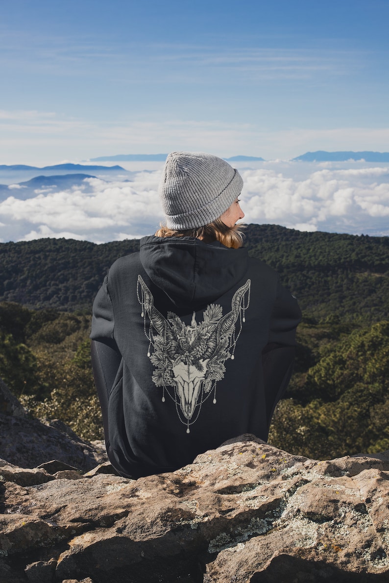 May include: A black hoodie with a detailed graphic of a skull with antlers, pine needles, and decorative chains. The person is wearing a gray beanie and is sitting on a rock with a scenic mountain backdrop.
