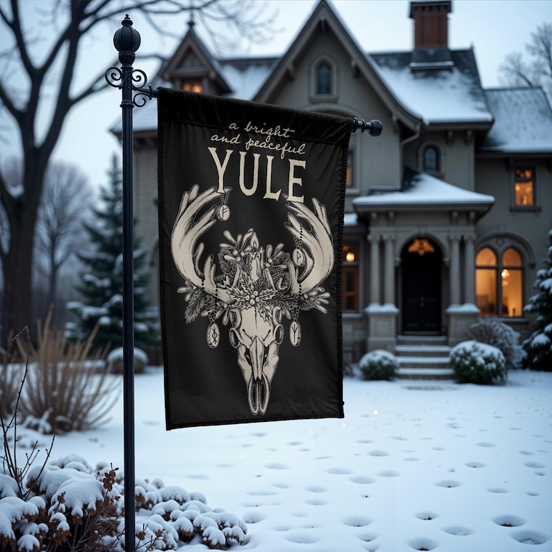 May include: A black flag with a white illustration of a deer skull with antlers adorned with ornaments and the text "a bright and peaceful YULE". The flag is hanging from a pole in front of a house covered in snow.