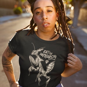 Może przedstawiać: Czarny T-shirt z grafiką dw&oacute;ch modliszek i tekstem "Eat the Patriarchy" z symbolem żeńskim.