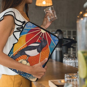 May include: A colorful laptop sleeve with a black and white magpie illustration. The sleeve features a geometric design with a sun, clouds, and a moon.