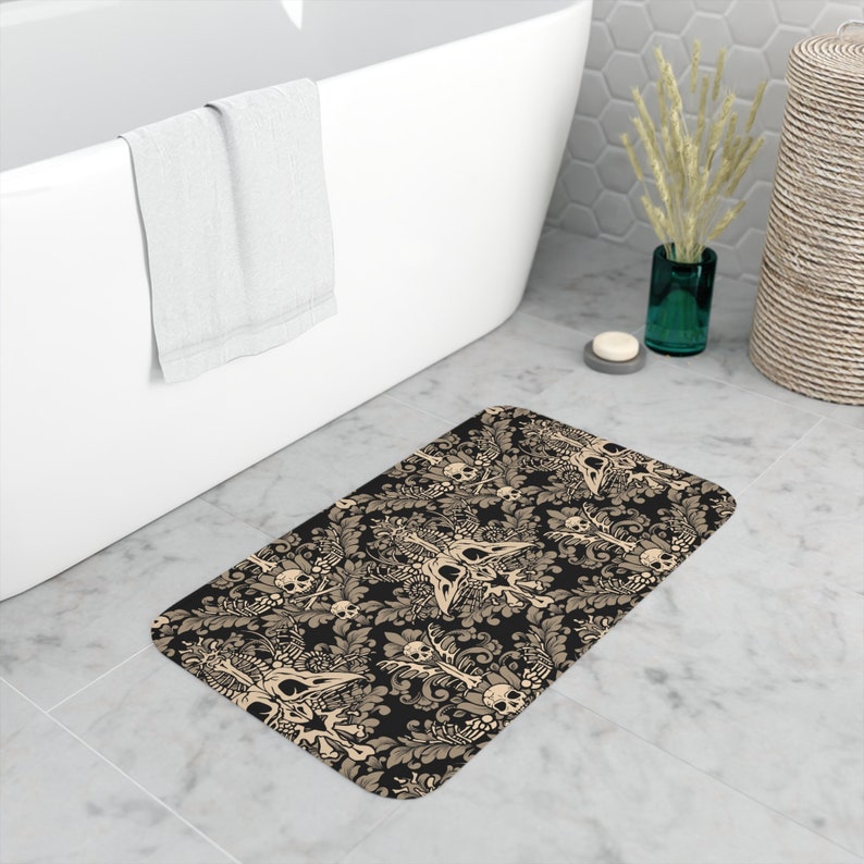 May include: A black and beige bath mat with a skull and crossbones pattern. The pattern is a damask design with skulls and crossbones in the center of each motif.