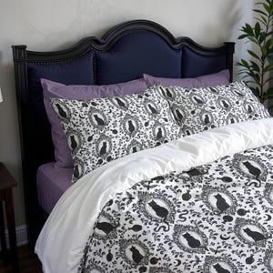 May include: A bed with a dark blue headboard and purple pillows. The bedding features a black and white pattern of framed black cat silhouettes, moons, and decorative swirls. A white duvet cover is folded over the patterned comforter.