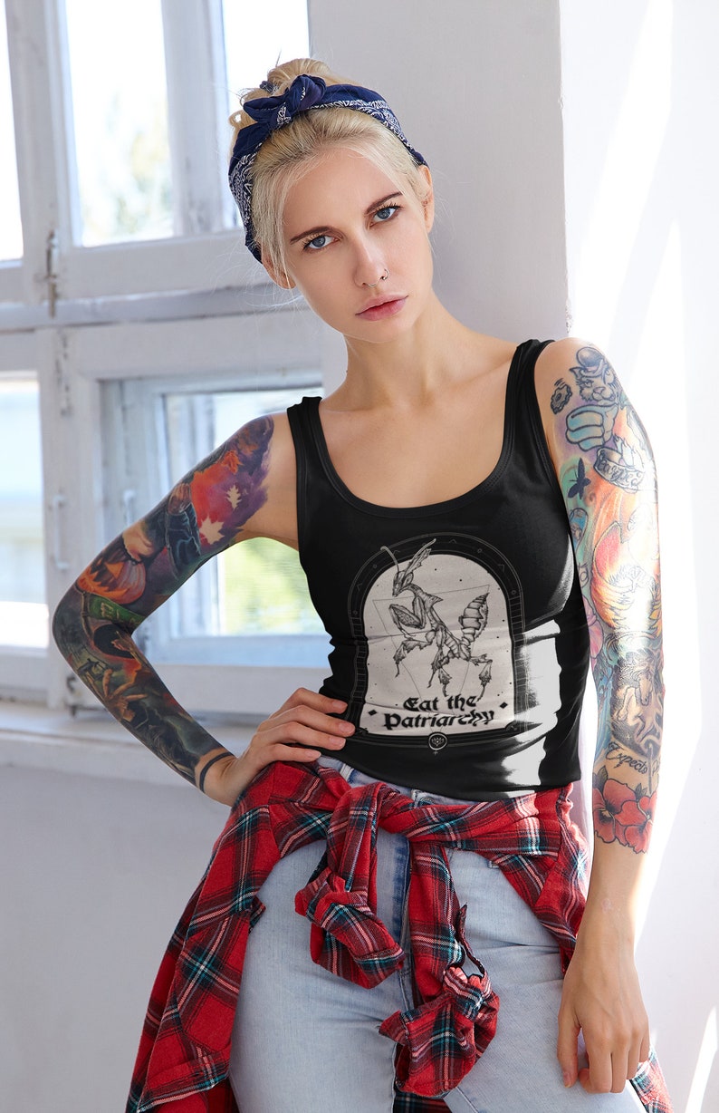 Może przedstawiać: Kobieta w czarnym topie na ramiączkach z grafiką modliszki i napisem "Eat the Patriarchy". Ma na sobie r&oacute;wnież czerwoną i czarną koszulę w kratę zawiązaną w talii i niebieskie dżinsy. Ma niebieską chustkę zawiązaną na głowie i tatuaże na ramionach.