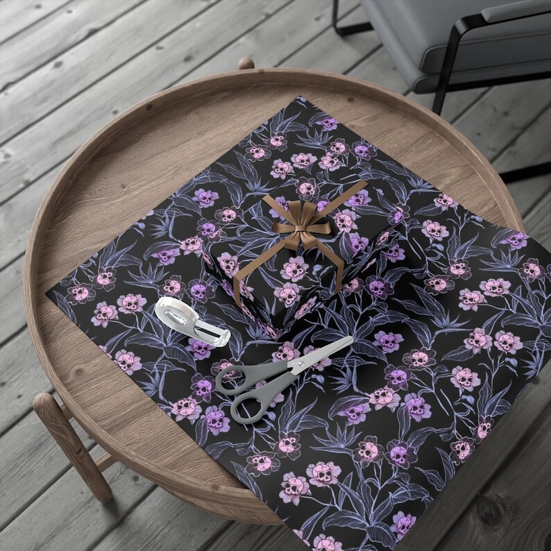 gothic floral gift wrapping paper with pastel skulls on a table with scissors, tape, and a wrapped present with a brown ribbon