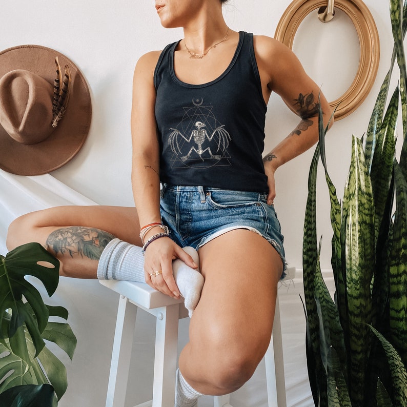 May include: A woman wearing a black tank top with a graphic of a skeletal bat inside a geometric design. The tank top has a scoop neck and thin straps. She is also wearing denim shorts and white socks.