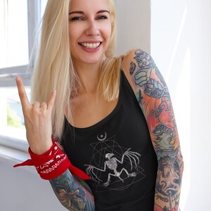 May include: A black tank top with a white graphic of a bat skeleton inside a geometric design. The tank top is being worn by a person with tattoos on their arms. The person is wearing a red bandana on their wrist and is making a rock and roll hand gesture.