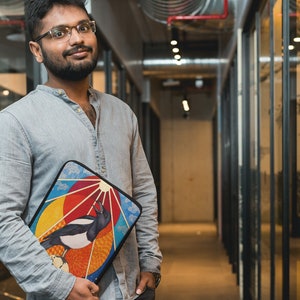 May include: A man wearing a light blue shirt and glasses is holding a laptop sleeve with a colorful design featuring a bird and a sun. The sleeve has a black background and a multi-colored design with a bird, a sun, and clouds.