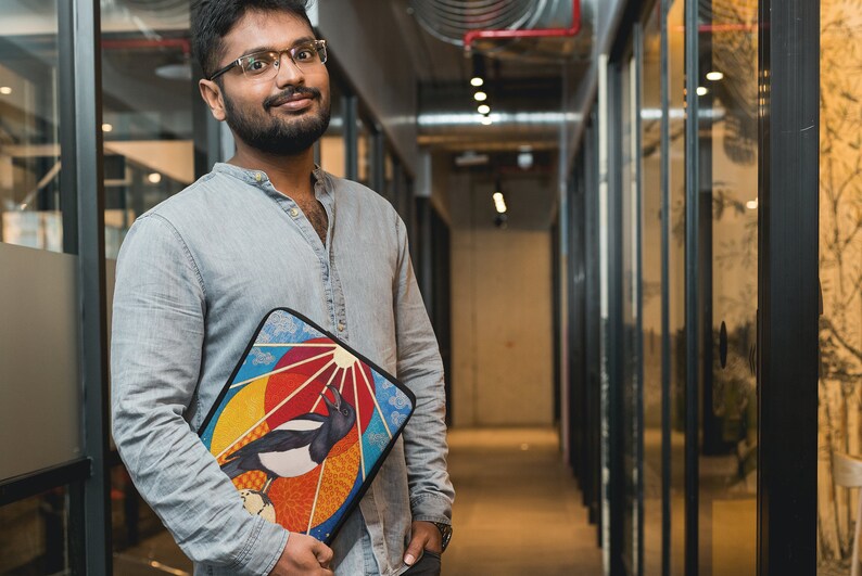 May include: A man wearing a light blue shirt and glasses is holding a laptop sleeve with a colorful design featuring a bird and a sun. The sleeve has a black background and a multi-colored design with a bird, a sun, and clouds.