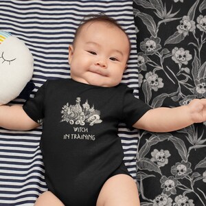 May include: A baby wearing a black bodysuit with a white graphic of a witch's cauldron and the text "Witch in Training".