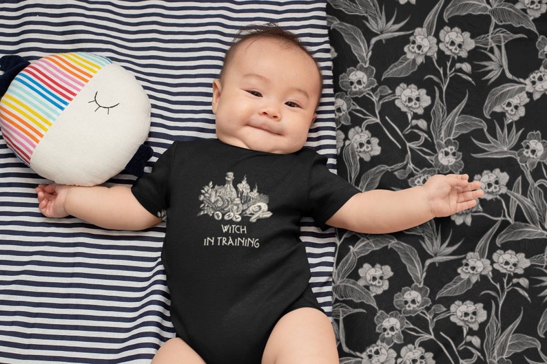 May include: A baby wearing a black bodysuit with a white graphic of a witch's cauldron and the text "Witch in Training".
