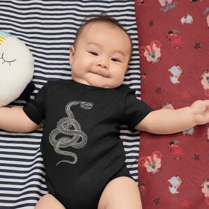 May include: A baby wearing a black bodysuit with a white snake graphic. The baby is lying on a striped blue and white blanket and a red blanket with a pattern of white goats.