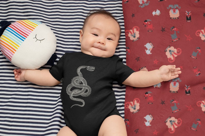 May include: A baby wearing a black bodysuit with a white snake graphic. The baby is lying on a striped blue and white blanket and a red blanket with a pattern of white goats.