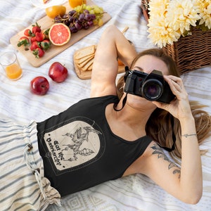 Może przedstawiać: Czarny top na ramiączkach z grafiką stworzenia z tekstem "Eat the Patriarchy." Top jest noszony przez osobę leżącą na białym i brązowym kocu z koszem piknikowym i jedzeniem.