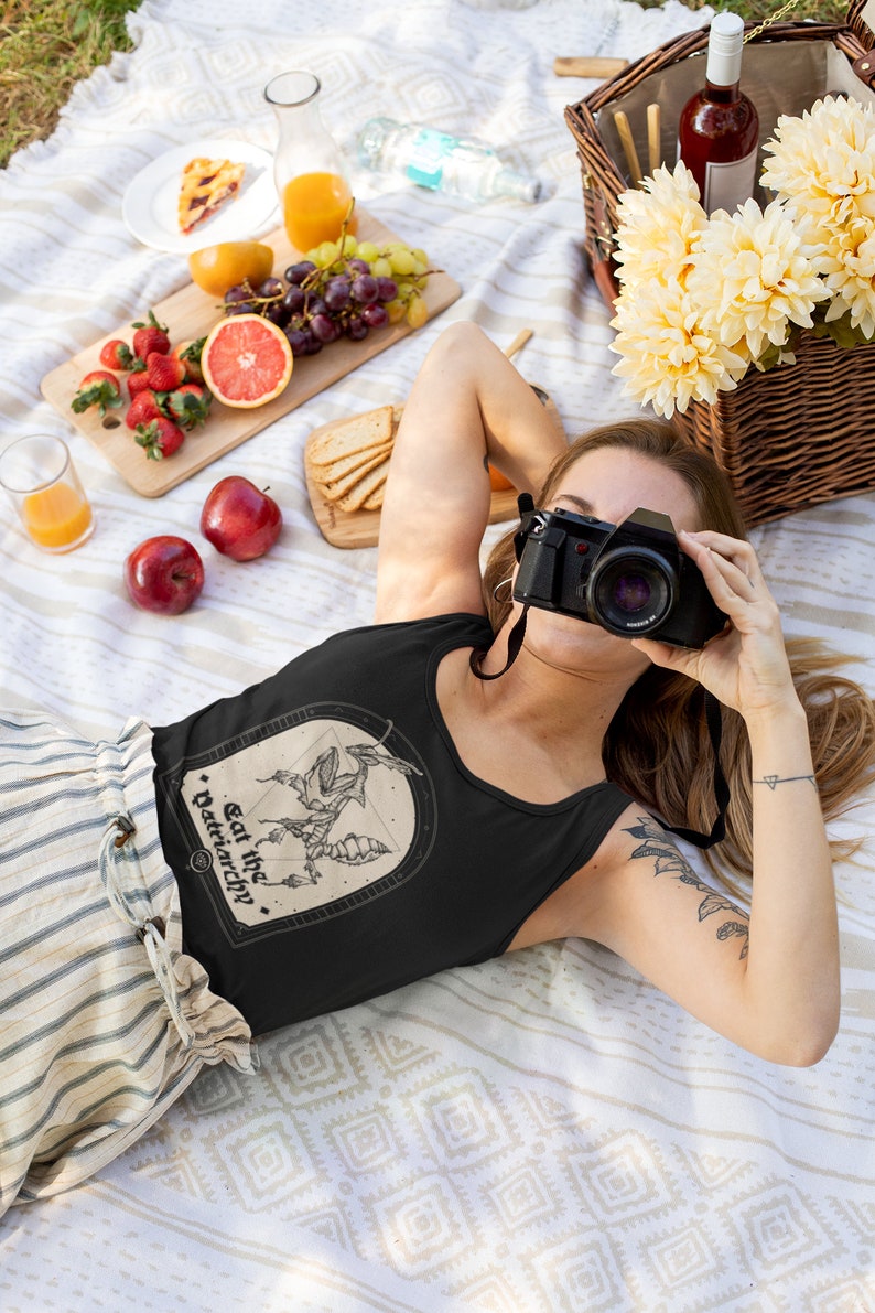 Może przedstawiać: Czarny top na ramiączkach z grafiką stworzenia z tekstem "Eat the Patriarchy." Top jest noszony przez osobę leżącą na białym i brązowym kocu z koszem piknikowym i jedzeniem.