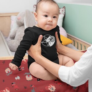 May include: A baby wearing a black long-sleeved onesie with a white pentagram and goat head design. The baby is sitting on a red blanket with a repeating pattern of goats and pentagrams.