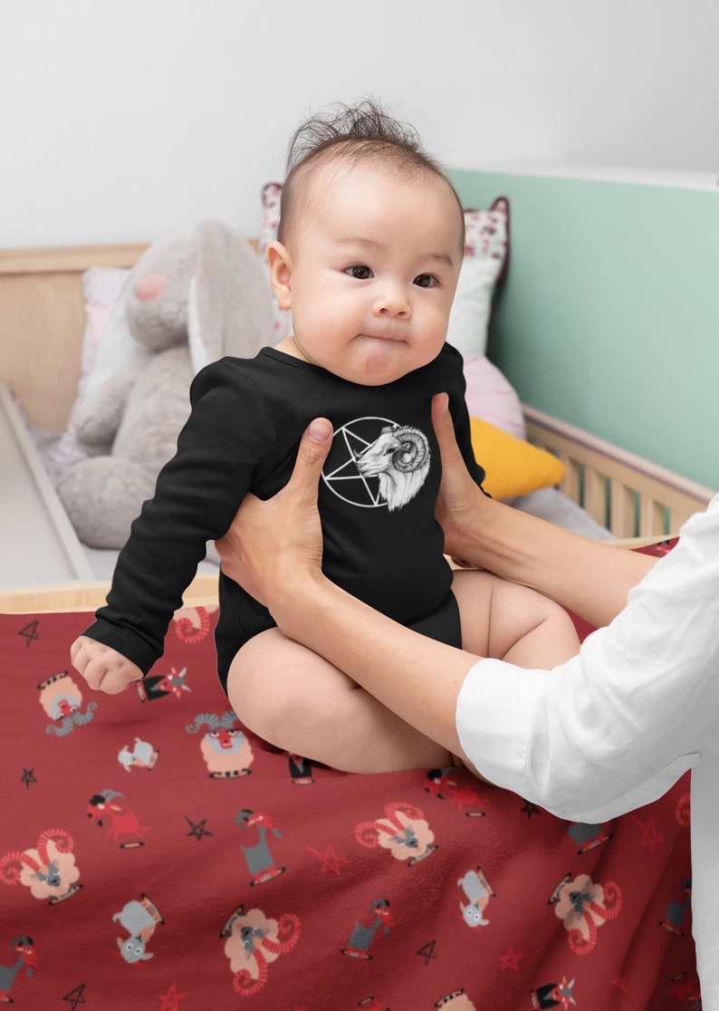 May include: A baby wearing a black long-sleeved onesie with a white pentagram and goat head design. The baby is sitting on a red blanket with a repeating pattern of goats and pentagrams.